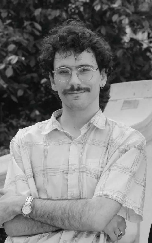 A greyscale image of Angelos Manolias, one of the creators and presenters of the DNA& podcast based on genetics. He is stood with his arms folded outside in a shirt.